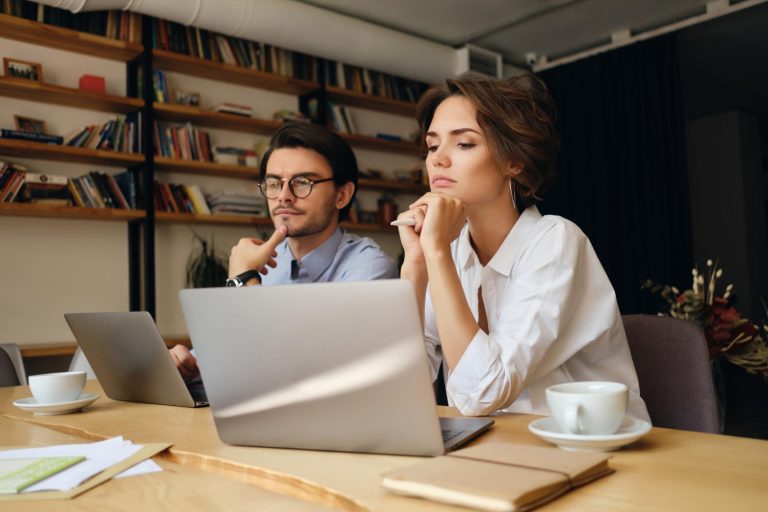 young-pensive-business-colleagues-sitting-desk-while-thoughtfully-working-laptop-with-coffee-modern-office